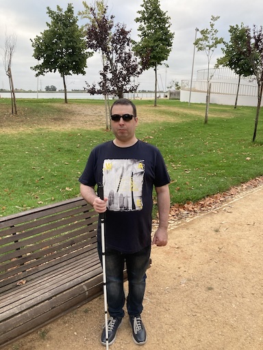 Me in T-shirt and pants wearing sunglasses and holding my white cane with some pruned bushes and overcast sky in the background